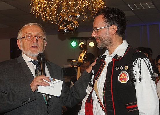 Zwei Männer stehen in einem Saal mit einem Mikrofon. Ein Mann hält ein Stück Papier in der Hand und spricht. Hinter ihnen sitzen Menschen, und ein Musikinstrument ist sichtbar. Ein Kronleuchter und grüne Lichter sind über ihnen.