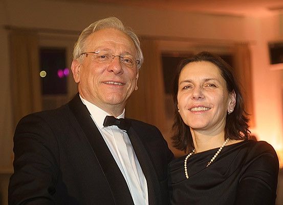 Ein Mann und eine Frau, beide lächelnd, stehen bei einer Veranstaltung nebeneinander. Der Mann trägt einen Frack mit einer Schleife und die Frau eine Perlenkette.