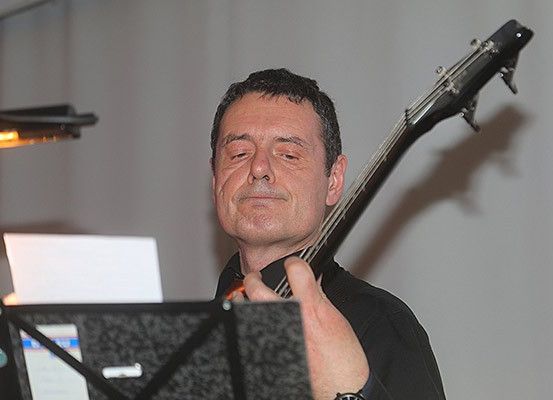 Ein Mann mit kurzem Haar spielt eine E-Gitarre vor einem Notenständer. Er trägt ein schwarzes Hemd und eine Uhr.