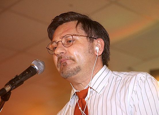 Ein Mann mit Brille und roter Krawatte steht vor einem Mikrofon und hält wahrscheinlich eine Rede.