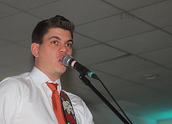 Ein Mann singt in ein Mikrofon, trägt ein weißes Hemd und eine rote Krawatte mit Totenkopfmuster. Die Umgebung scheint ein Indoor-Event zu sein.