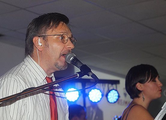 Ein Mann singt in ein Mikrofon auf der Bühne, begleitet von einer Frau mit kurzen Haaren, die in ein weiteres Mikrofon singt. Die Bühne ist mit blauen Bühnenlichtern beleuchtet.