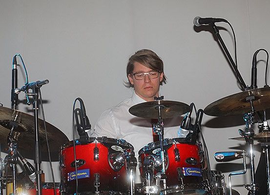 Ein Mann spielt die Drums bei einer Aufführung, mit einem weißen Hemd, Brille und umgeben von einem Schlagzeug, Mikrofonen und Becken.
