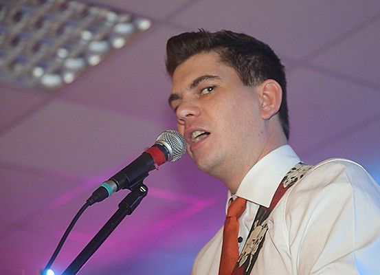 Ein Mann singt in ein Mikrofon, auf das ein Scheinwerfer gerichtet ist. Er trägt ein weißes Hemd, eine rote Krawatte und gemusterte Hosenträger. Das Mikrofon ist schwarz und rot.