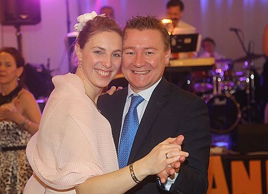 Ein Mann und eine Frau tanzen eng bei einer Party, mit einer Band im Hintergrund. Der Mann trägt einen Anzug und eine Krawatte, und die Frau trägt ein rosa Kleid.