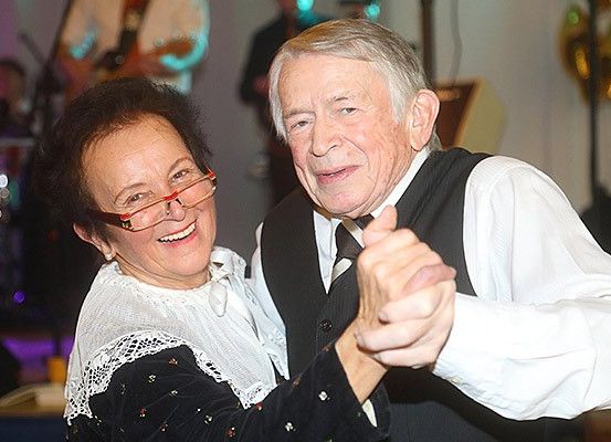 Ein älteres Paar tanzt in einer Location, lächelt und hält sich aneinander. Die Frau trägt eine Brille und einen weißen Spitzenschal. Hinter ihnen spielen Musiker Gitarre und Schlagzeug.