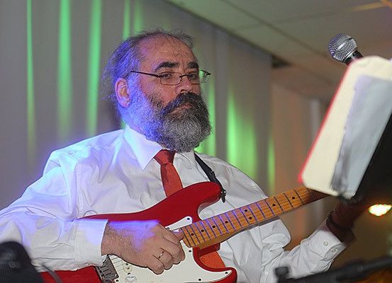 Ein älterer Mann mit Bart spielt eine rote E-Gitarre. Er trägt ein weißes Hemd, eine rote Krawatte und eine Brille. Ein Mikrofon und ein Notenständer stehen vor ihm. Der Hintergrund hat grüne Lichter.