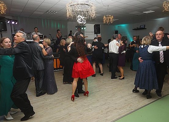 Eine Gruppe von Menschen tanzt in einem Ballsaal, wobei eine Kronleuchter und Lautsprecher sichtbar sind. Die Tänzer bestehen aus Männern und Frauen in formeller Kleidung, und eine Bühne mit einer Band ist im Hintergrund sichtbar.