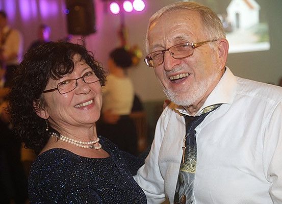 Ein älteres Paar, beide mit Brille, lächelt und steht eng beieinander auf einer Party. Sie sind von anderen tanzenden Menschen umgeben. Der Mann trägt eine Krawatte.
