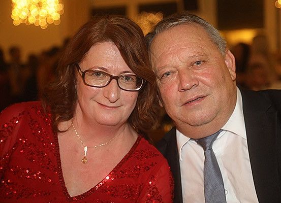 Ein Mann und eine Frau in formeller Kleidung lächeln für die Kamera bei einer Veranstaltung. Die Frau trägt eine Brille und eine Halskette.