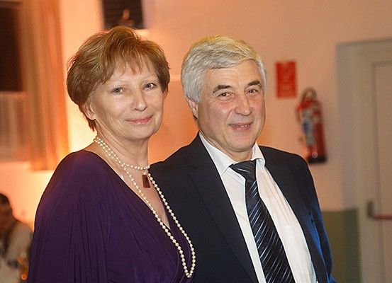 Ein Mann und eine Frau stehen eng beieinander und lächeln für ein Foto in einer formellen Umgebung. Der Mann ist in Anzug und Krawatte gekleidet, während die Frau eine Halskette und eine lila Bluse trägt. Im Hintergrund befindet sich ein Feuerlöscher und eine Tür.