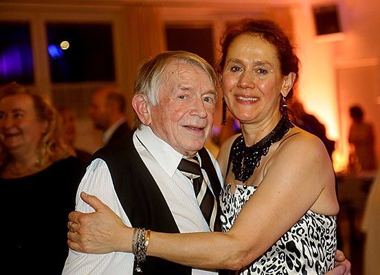 Ein älterer Mann und eine Frau umarmen sich bei einer Party und lächeln. Im Hintergrund sind andere Leute zu sehen.