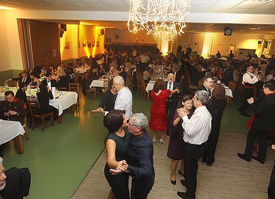 Eine große Gruppe von Menschen versammelt sich für eine formelle Veranstaltung und tanzt in der Mitte eines Raumes, um den herum Tische und Stühle aufgestellt sind.