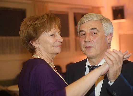 Ein älteres Paar tanzt bei einer formellen Veranstaltung. Die Frau trägt ein lilafarbenes Oberteil und eine Perlenkette, während der Mann einen Anzug und eine Krawatte trägt. Sie lächeln sich mit warmen Gesichtsausdrücken an.