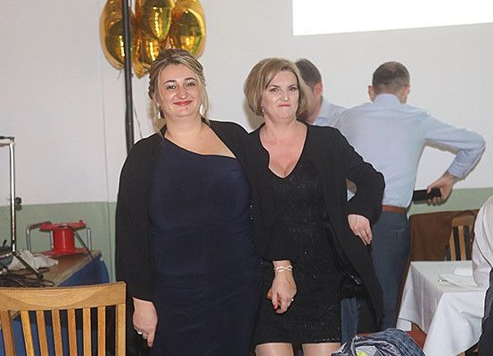 Zwei Frauen stehen eng beieinander in einem Raum mit Luftballons und Menschen, die an Tischen sitzen. Ein Mann hält ein Handy hinter ihnen.