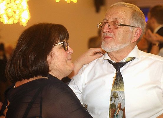 Ein älterer Mann mit weißem Bart lässt sich von einer älteren Frau den Krawattenknoten zurechtrücken. Sie tragen beide eine Brille und sind von anderen Gästen auf einer Party umgeben.