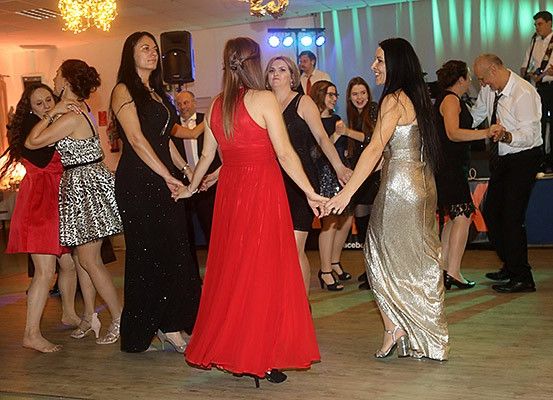 Eine Gruppe von Frauen in formeller Kleidung tanzt auf einem Holzboden. Eine Frau in einem roten Kleid hält die Hände von zwei anderen. Hinter ihnen steht ein Mann, der eine Gitarre spielt.