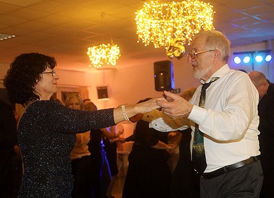 Ein älteres Paar tanzt in einem Raum mit anderen Leuten, Kronleuchtern und Disco-Lichtern.