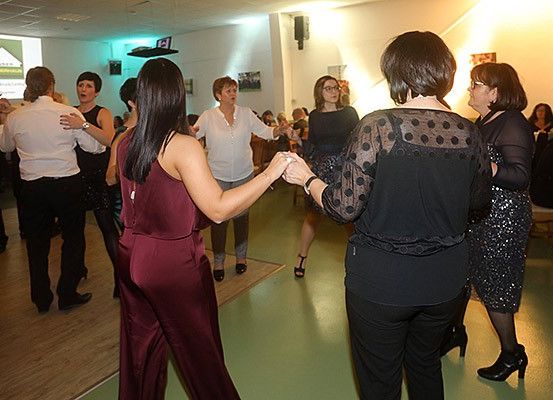 Drinnen tanzen eine Gruppe von Erwachsenen, darunter Frauen und Männer, während eine Frau sie anführt. Eine Projektionsleinwand ist an der Wand montiert und ein Projektor befindet sich an der Decke.