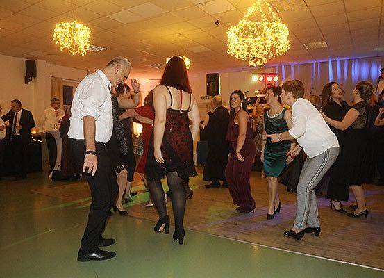 Eine Gruppe von Menschen tanzt in einem Raum mit Kronleuchtern und einem Holzboden. Einige tragen formelle Kleidung.