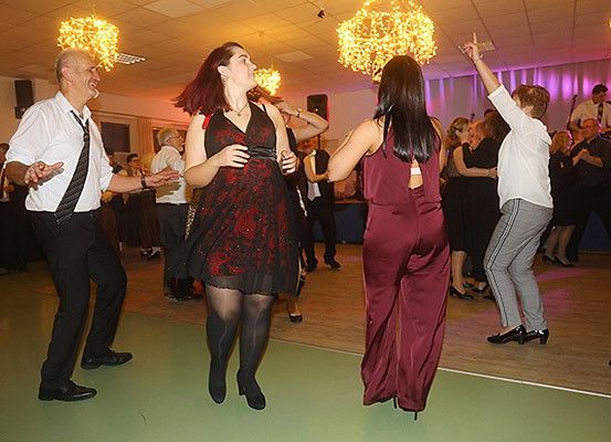 Eine Gruppe von Menschen tanzt auf einer Party. Die Frau in der Mitte trägt ein rotes Kleid und schwarze Strümpfe. Der Mann links trägt ein weißes Hemd und eine Krawatte.