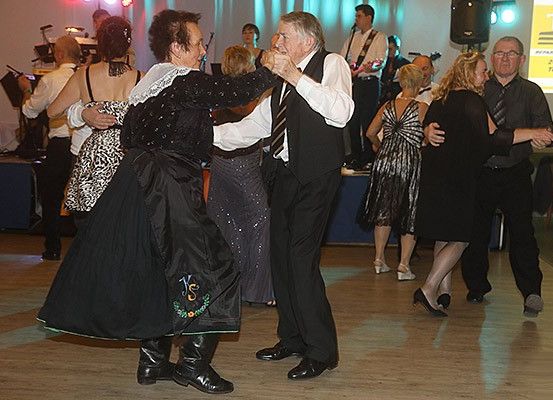Eine Gruppe von Menschen tanzt auf einem Holzboden, wobei ein Mann und eine Frau in der Mitte schwarze Kleider und Stiefel tragen. Hinter ihnen tanzen andere Paare, während eine Band im Hintergrund spielt.