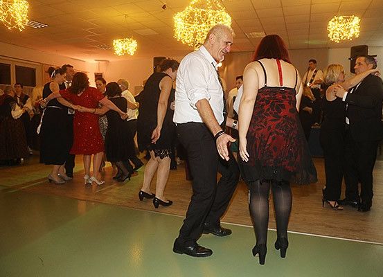 Eine Gruppe von Menschen tanzt auf einer Party, einige in formeller Kleidung. Ein Mann und eine Frau tanzen eng zusammen. Sie befinden sich in einem Raum mit Holzboden und dekorativen Lichtern.