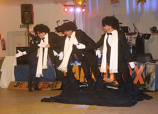 Drei Männer in Anzügen und weißen Handschuhen treten auf der Bühne auf, mit einem Hintergrund und Musikinstrumenten im Hintergrund.