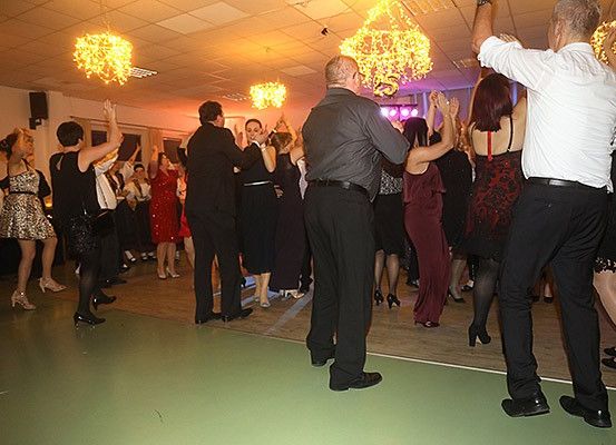 Eine Gruppe von Menschen tanzt in einem Partysaal mit Kronleuchtern. Einige tragen hohe Absätze und andere tragen Lederschuhe.
