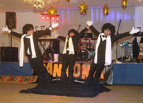 Drei Männer in Anzügen treten auf einer Bühne auf, umgeben von Musikinstrumenten, Luftballons und einem Banner mit orangen Buchstaben.