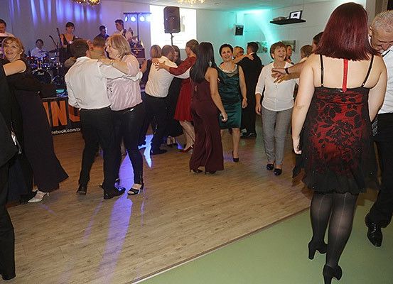 Menschen tanzen in einem Raum mit Holzboden und lila Beleuchtung. Eine Band spielt im Hintergrund.