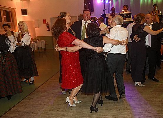 Eine Gruppe von Menschen tanzt in einem Raum mit Bühne, einige in formeller Kleidung und andere in traditioneller Tracht.