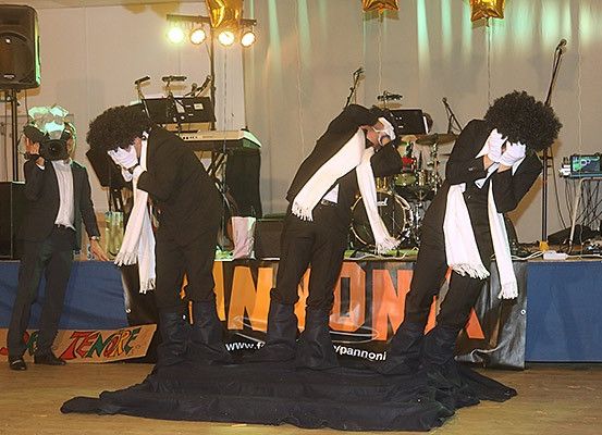 Eine Gruppe von Darstellern mit Masken auf der Bühne. Sie sind in schwarzen Anzügen und weißen Schals gekleidet. Die Bühne hat einen schwarzen Stoff, ein Schlagzeug und Musikinstrumente. Dahinter steht ein Tisch, der mit einem blauen Tuch bedeckt ist.