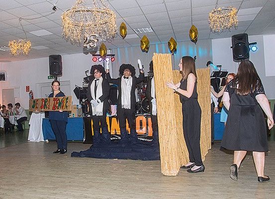 Mehrere Personen stehen auf der Bühne, zwei Männer in Anzügen und Perücken halten Schilder. Dahinter befindet sich ein Banner und Lautsprecher. Eine Frau hält ein Schild. Im Hintergrund sitzen Leute an Tischen.