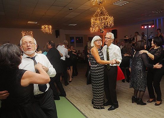 Eine Gruppe älterer Menschen tanzt in einem Ballsaal, mit Kronleuchtern, die von der Decke hängen. Ein Mann und eine Frau umarmen sich.