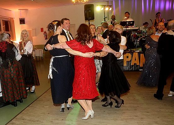 Mehrere Menschen tanzen in einem Festsaal. Eine Band spielt hinter ihnen. Ein Tisch steht links und ein Banner rechts. Ein Feuerlöscher ist an der Wand montiert.