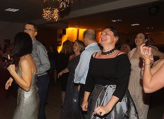 Mehrere Menschen tanzen auf einer Party. Eine Frau in einem silbernen Kleid ist im Vordergrund. Hinter ihr lächelt eine Frau in einem schwarzen Kleid und einer Halskette. Ein älterer Mann mit Brille steht hinter ihr. Andere Gäste tanzen im Hintergrund.