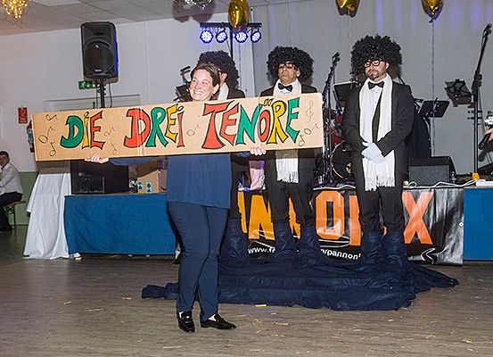 Eine Frau hält ein Schild mit der Aufschrift Die Drei Tenore, während zwei Männer in Anzügen hinter ihr stehen. Sie befinden sich auf einer Bühne, und Musikequipment ist im Hintergrund zu sehen.