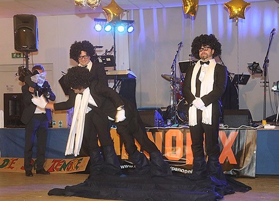 Vier Männer in Anzügen und Perücken treten mit Musikinstrumenten, Lautsprechern und Luftballons im Hintergrund auf der Bühne auf.