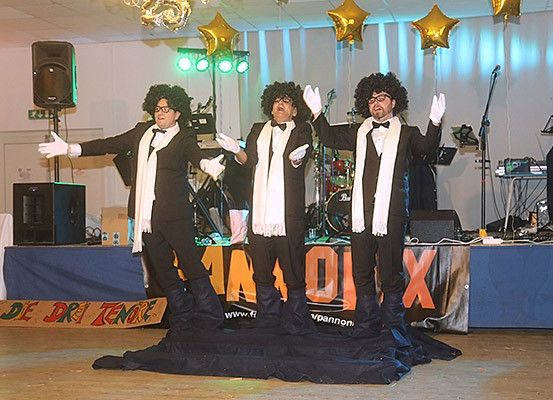 Drei Männer in Smokings stehen auf der Bühne und führen möglicherweise einen musikalischen Auftritt durch. Sie tragen weiße Handschuhe und Fliegen. Hinter ihnen befinden sich Musikinstrumente, Lautsprecher und ein Banner mit der Aufschrift 'MA OI X'.