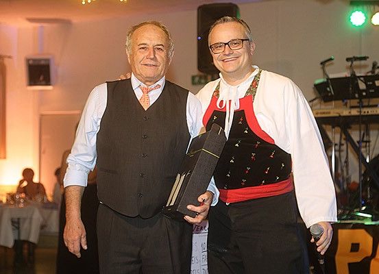 Zwei Männer stehen nebeneinander, einer hält eine Trophäe und lächelt, während der andere ein Mikrofon hält. Sie befinden sich in einem Raum mit einer musikalischen Einrichtung, einem Banner und Menschen, die an Tischen im Hintergrund sitzen.