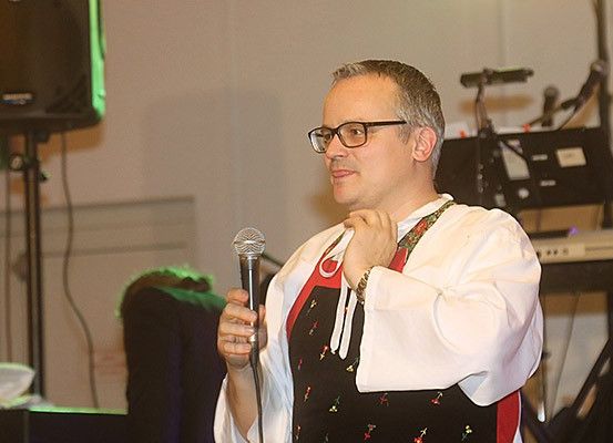 Ein Mann mit Brille und einem weißen Hemd mit schwarzem Schürze hält ein Mikrofon und spricht. Dahinter steht ein schwarzer Lautsprecher, ein Klavier und eine Person sitzt auf einem Stuhl.