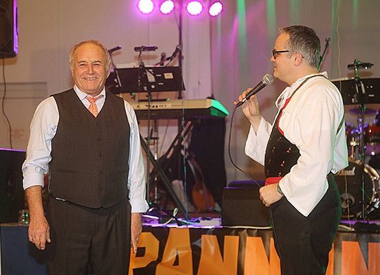 Zwei Männer auf der Bühne, einer singt in ein Mikrofon. Die Bühne hat ein Banner mit Text, einen Tisch, Lautsprecher und Musikinstrumente.
