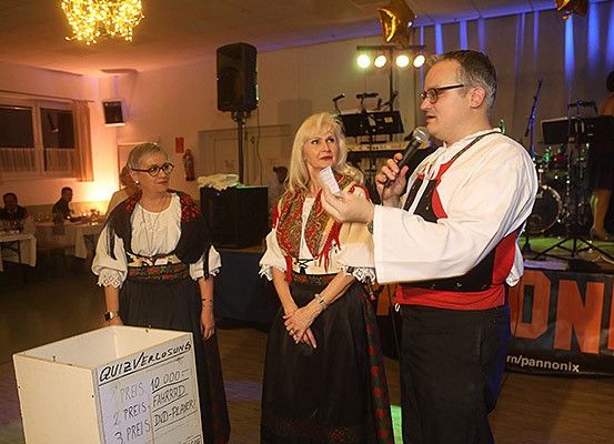 Zwei Frauen in traditioneller Kleidung stehen neben einem Podium, während ein Mann in ein Mikrofon spricht. Hinter ihnen steht ein Banner mit der Aufschrift 'ONI'. Der Raum ist mit Lichtern und einer Laterne dekoriert.