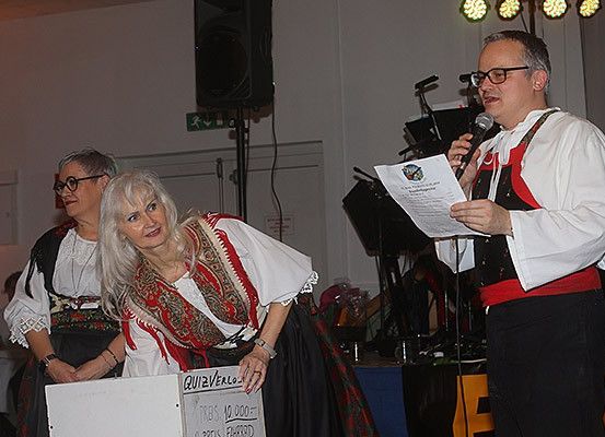 Ein Mann spricht in ein Mikrofon bei einer Veranstaltung, mit einer Frau neben ihm, die eine Box mit Text und Zahlen hält. Dahinter eine Frau in traditioneller Kleidung und ein Lautsprecher.