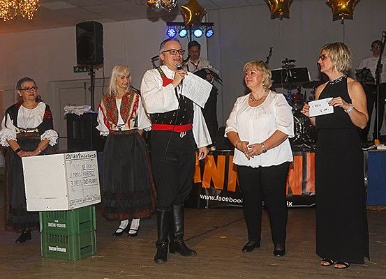 Mehrere Personen stehen auf einer Bühne, ein Mann spricht in ein Mikrofon. Frauen in traditionellen Kostümen und Abendkleidern sind anwesend. Ein Banner ist hinter ihnen sichtbar.