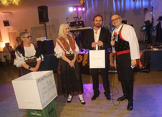 Drei Personen stehen auf einer Bühne und halten eine Tasche und ein Schild. Hinter ihnen steht eine Frau neben einer Kiste. Ein Mann spricht in ein Mikrofon. Dahinter befindet sich ein Tisch mit Musikinstrumenten und einem Lautsprecher.