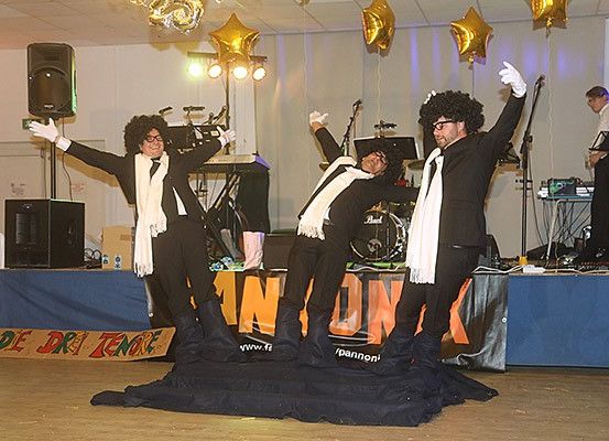 Drei Personen in schwarzen Anzügen und weißen Handschuhen stehen auf der Bühne und führen eine Performance mit einer Band durch. Ein Schlagzeug, Lautsprecher und eine Gitarre stehen hinter ihnen. Ein Banner mit der Aufschrift 'DE DRI TENORE' liegt auf dem Boden.