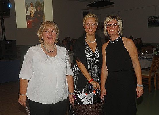 Drei Frauen stehen zusammen in einem Raum. Die Frau auf der linken Seite trägt eine weiße Bluse, die in der Mitte ein schwarzes Kleid mit einem Korb und die Frau auf der rechten Seite ebenfalls ein schwarzes Kleid. Hinter ihnen sind Stühle, Tische und eine Projektionsleinwand.