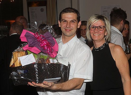 Ein Mann und eine Frau halten einen Geschenkkorb mit einer rosa Schleife und stehen in einem Raum. Die Frau trägt eine Brille. Hinter ihnen stehen mehrere Personen und schauen sie an.